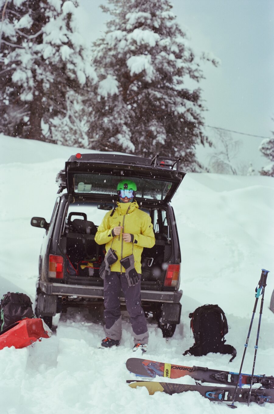 Rider in a yellow jacket gearing up at the open trunk of a 4x4 in deep snow