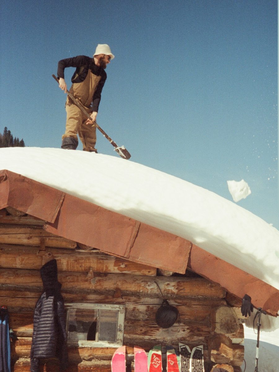 Shovelling snow off a wooden hut roof, skis lined up against the wall