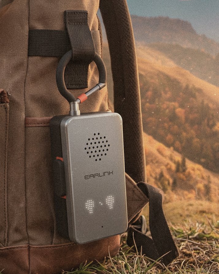 EarLink clipped to a backpack strap by its carabiner antenna, mountains behind
